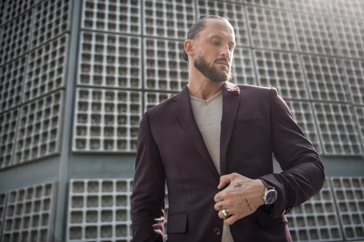 Mężczyzna w beżowym swetrze, bordowej marynarce z zegarkiem na ręku i pierścionkiem na palcu.