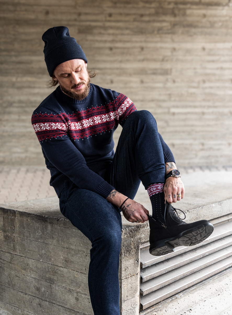 Mężczyzna ubrany w czarną czapkę, świąteczny sweter, ciemne jeansy.