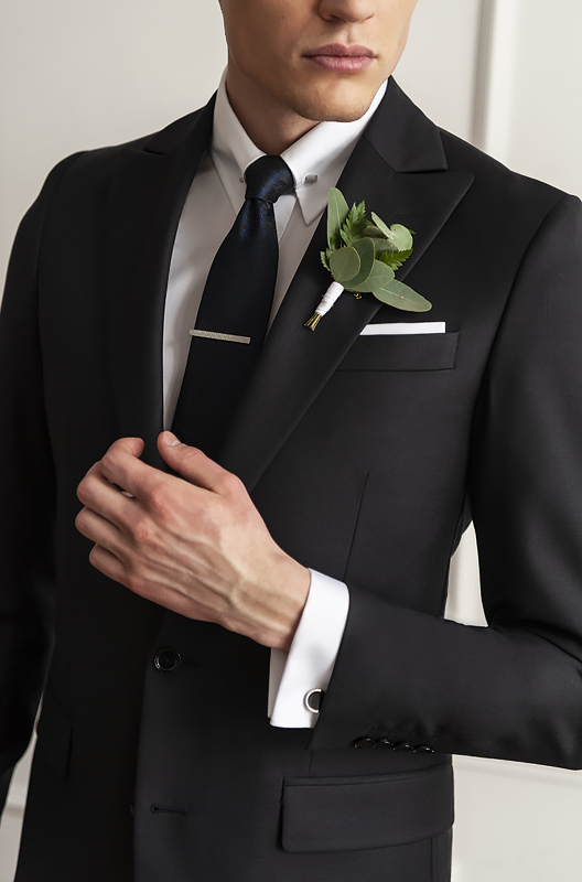 Model ubrany w czarny smoking, białą koszulę z czarnymi guzikami, czarny krawat z butonierką na klapie