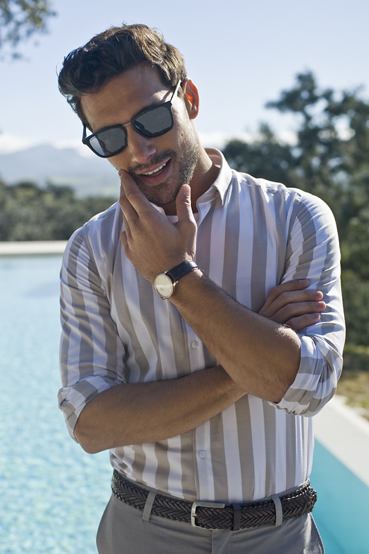 Mężczyzna ubrany w koszulę w beżowe paski, szare spodnie i pasek, z zegarkiem na ręku i w okularach na twarzy